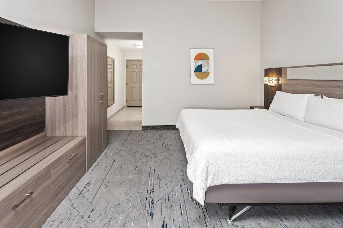 A hotel room with a large bed, a flat-screen TV mounted on the wall, and a hallway leading to another room.