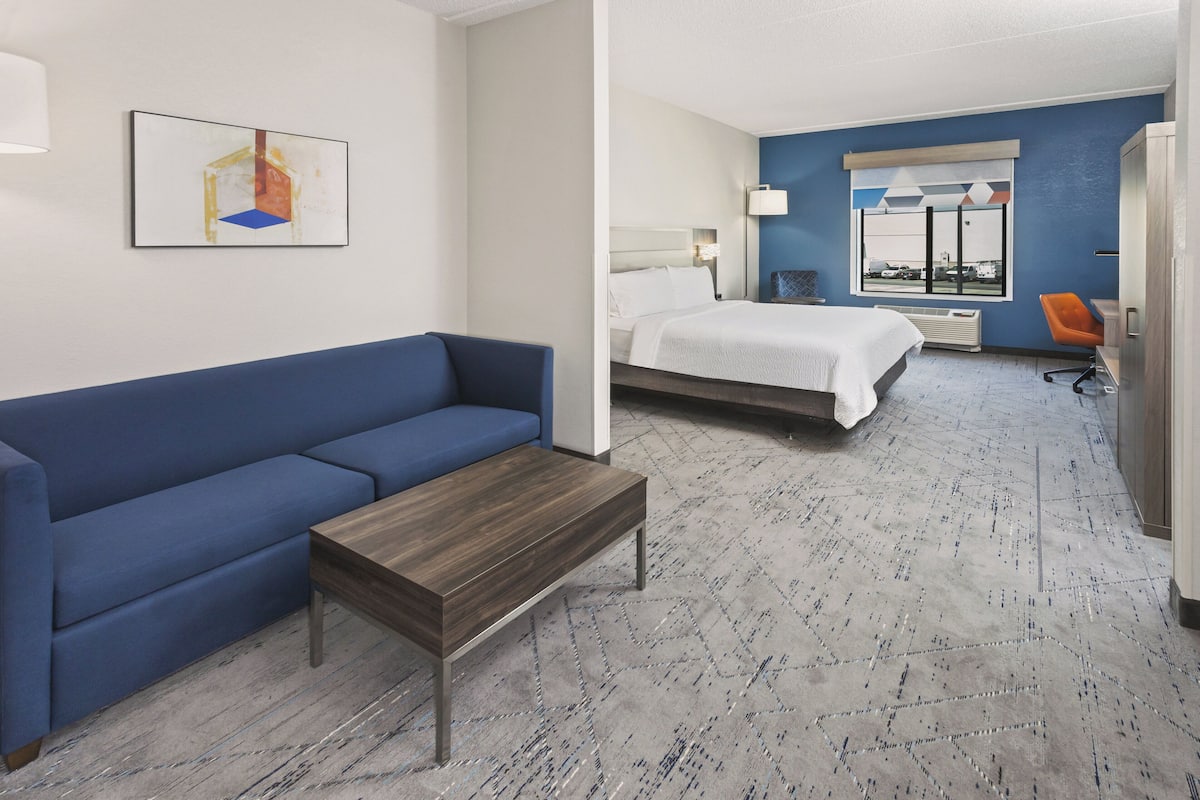 A hotel room with a blue sofa, a wooden coffee table, a bed with white bedding, a desk with a chair, and a window with a view.