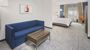 A hotel room with a blue sofa, a wooden coffee table, a bed with white linens, a desk with a chair, and a window with curtains.