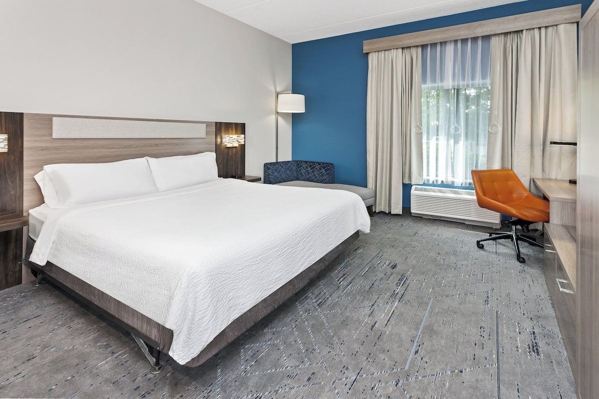 A hotel room with a large bed, a desk, and a chair. There is a window with curtains and a lamp on the desk.