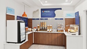 A breakfast area with a refrigerator, a coffee machine, and a display of fruits, cereals, and bakery items.