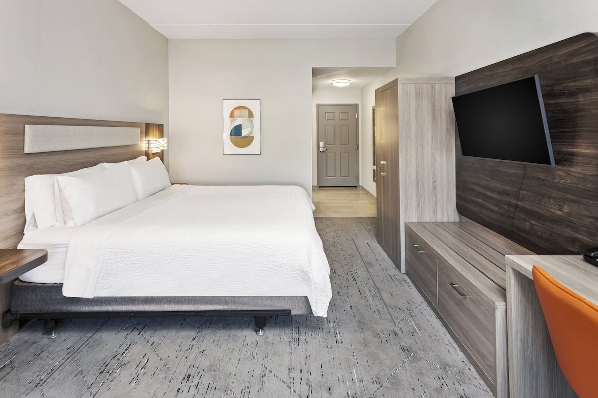 A hotel room with a large bed, a TV mounted on the wall, a desk with a chair, and a door leading to another room.