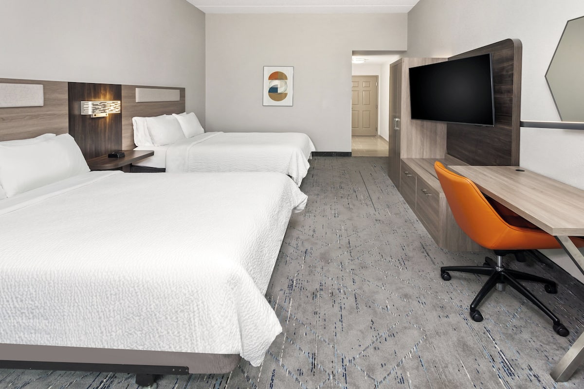 A hotel room with two beds, a desk with an orange chair, a flat-screen TV, and a wall-mounted artwork.