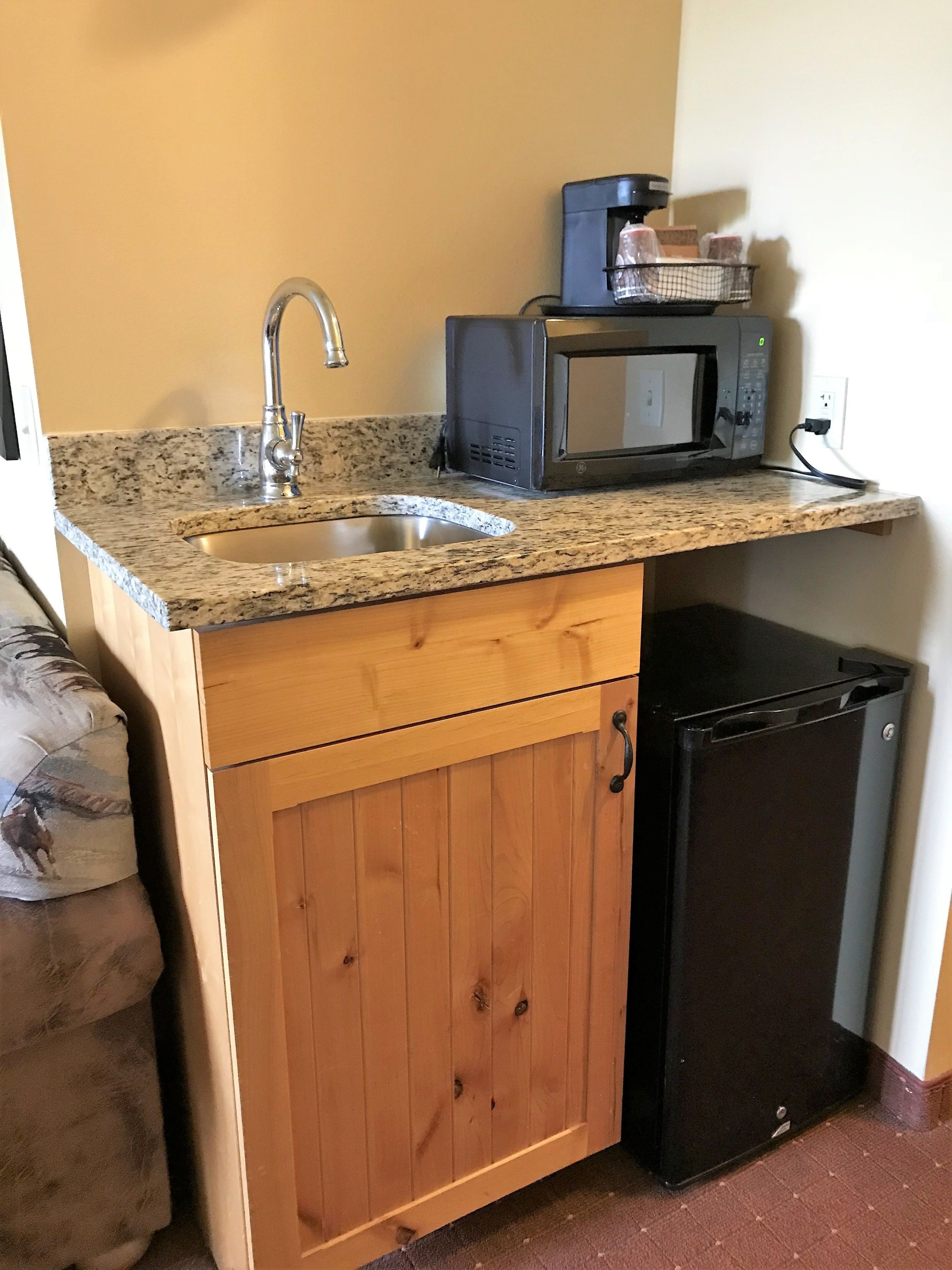 A compact kitchen with a granite countertop, stainless steel sink, a microwave, and a small black refrigerator.