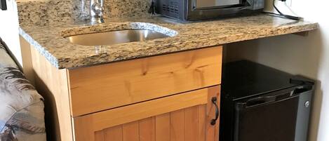 A compact kitchen with a granite countertop, stainless steel sink, a microwave, and a small black refrigerator.