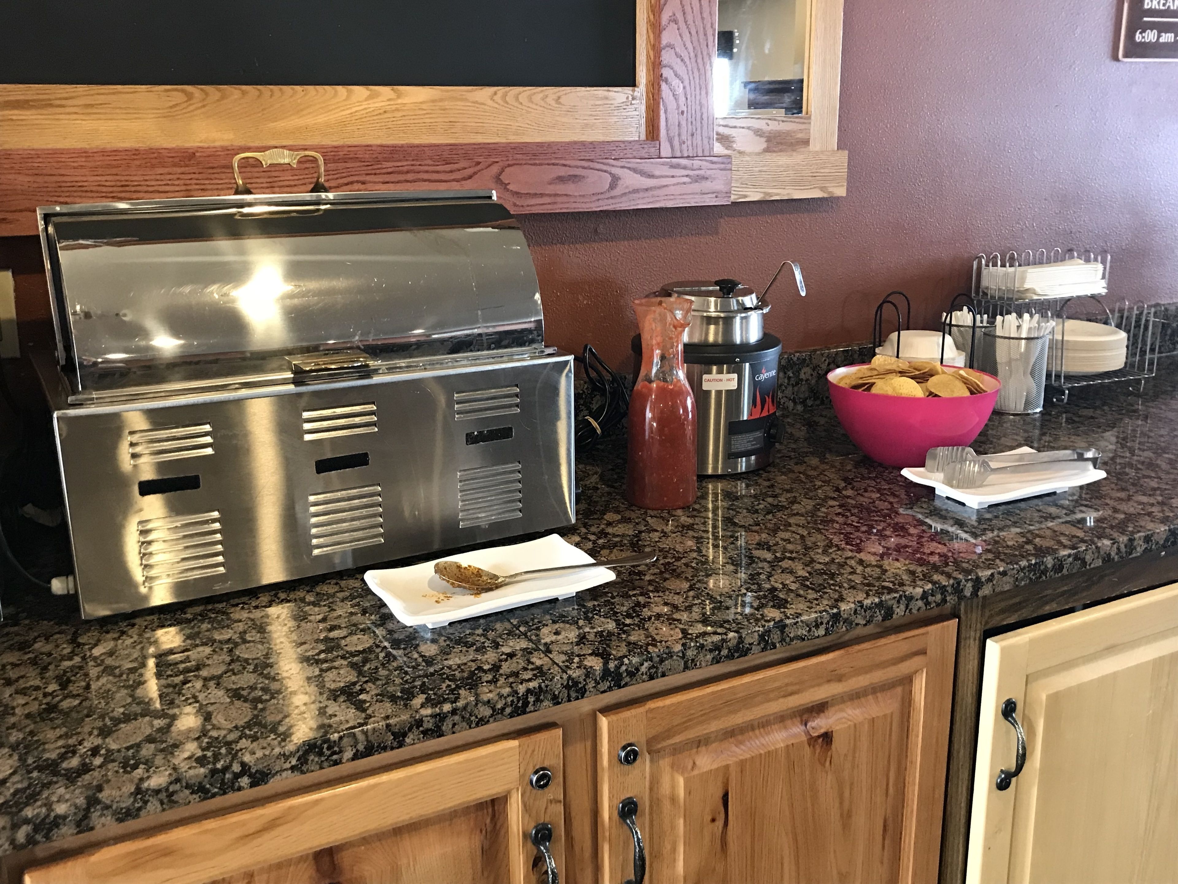 A kitchen counter with a stainless steel grill, a Cajun seasoning dispenser, a bottle of hot sauce, a bowl of tortilla chips, and a plate with a tortilla chip and a spoon.