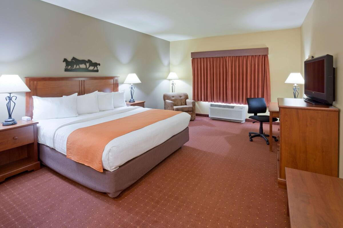 A hotel room with a large bed, two bedside tables with lamps, a desk with a chair, a television, and a large window with curtains.