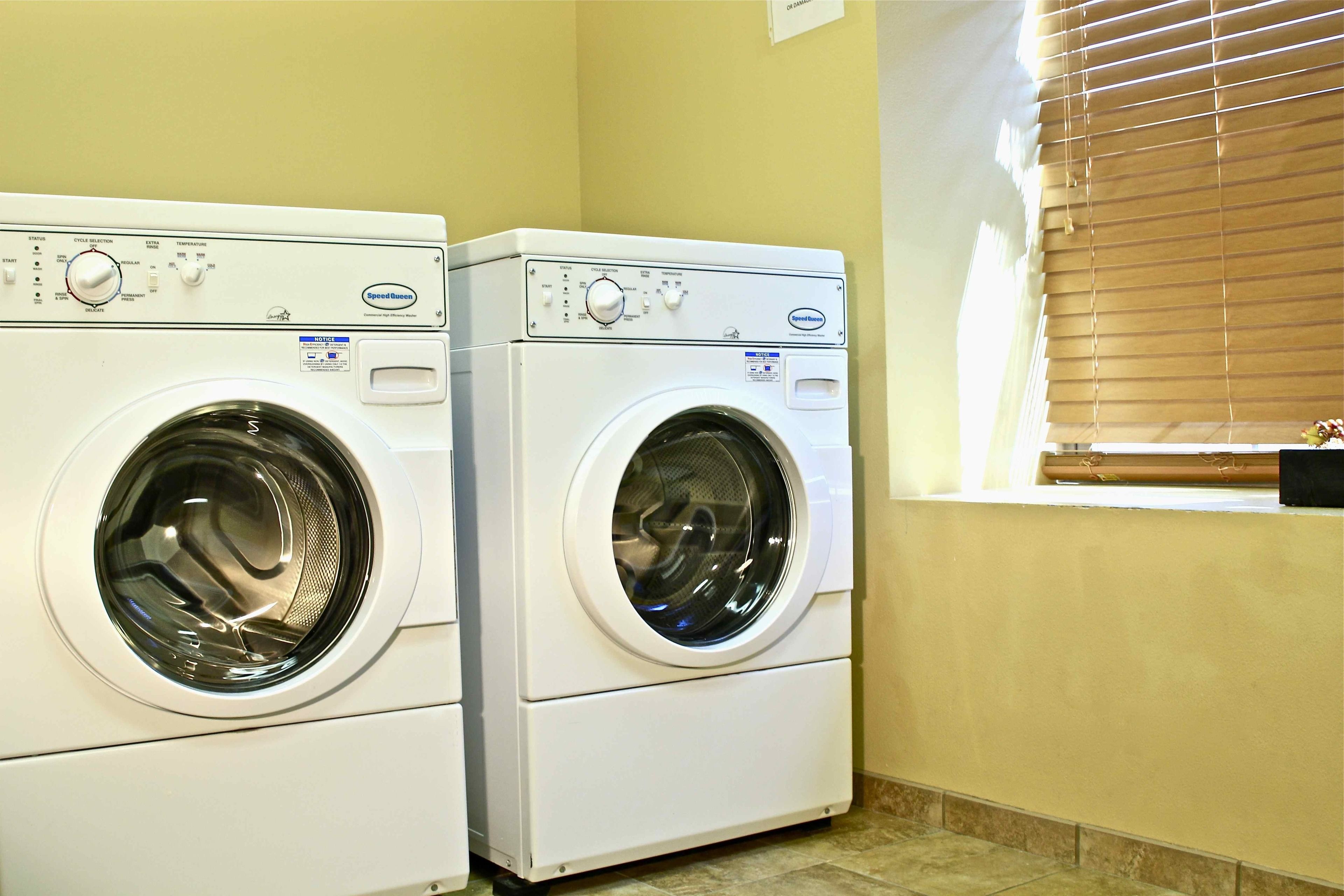 Two front-loading washing machines in a laundry room with a window and blinds.