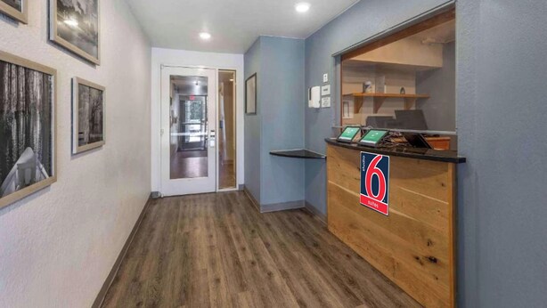 A hallway with wooden flooring, a reception desk, framed pictures on the wall, and a door leading to another room. - Studio 6 Extended Stay Shreveport Airport LA (Shreveport)
