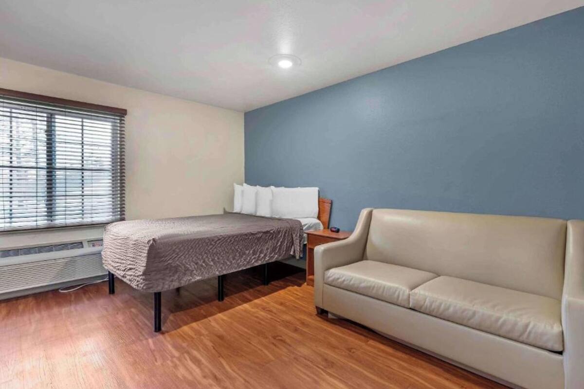 A bedroom with a bed, a nightstand, a sofa, a window with blinds, and a radiator.
