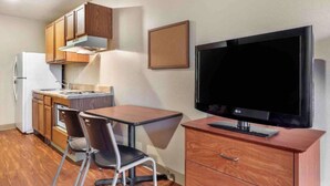 A compact kitchen with wooden cabinets, a white refrigerator, a small dining table with two chairs, and a flat-screen TV on a wooden stand. - Studio 6 Extended Stay Shreveport Airport LA (Shreveport)