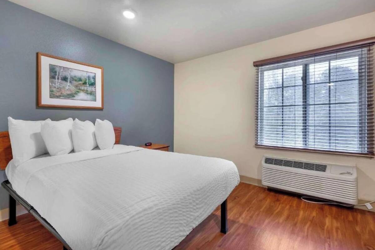 A bedroom with a bed, a framed picture on the wall, a window with blinds, and a wall-mounted air conditioner.