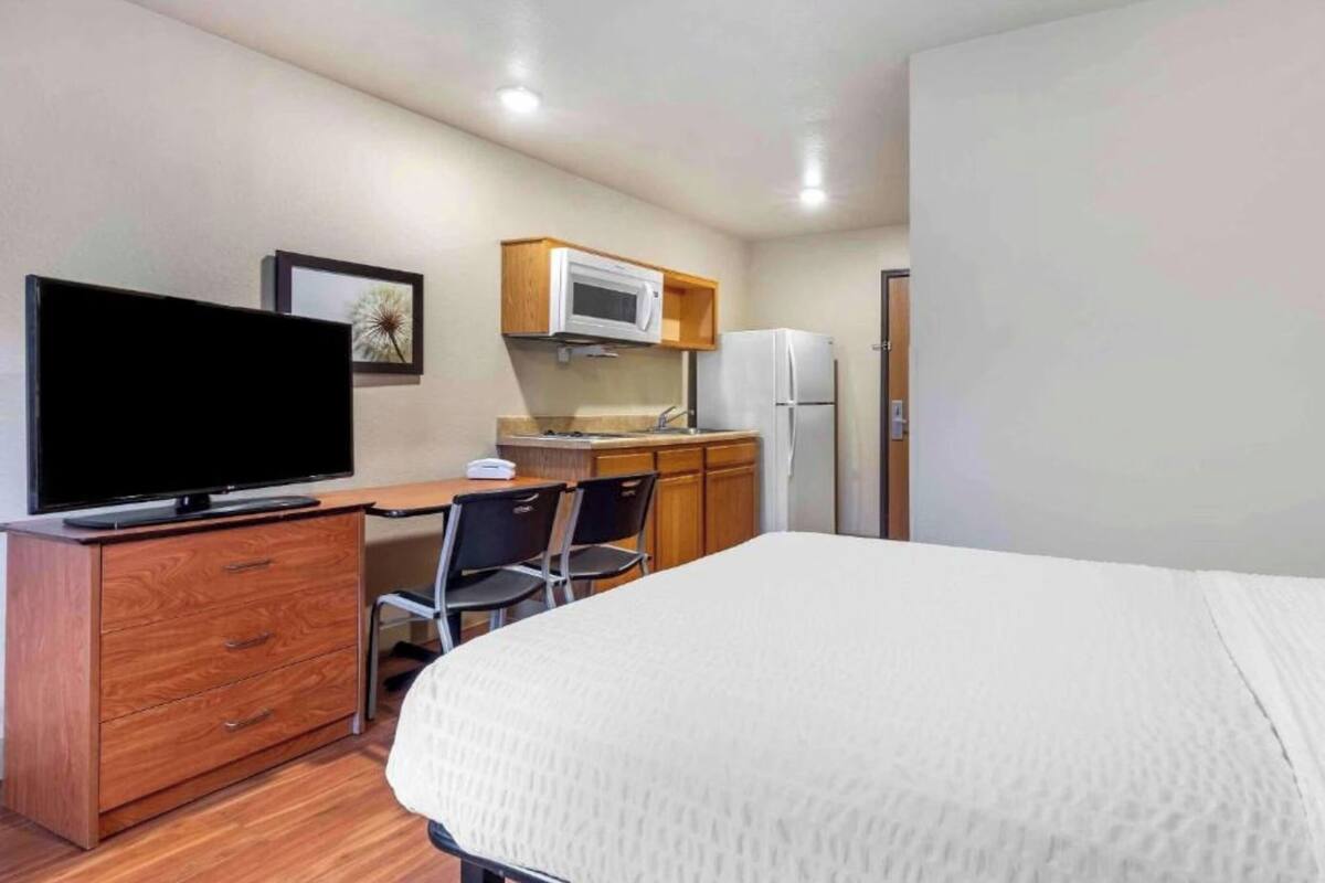 A hotel room with a bed, a TV on a wooden dresser, a kitchenette with a microwave and refrigerator, and a chair.