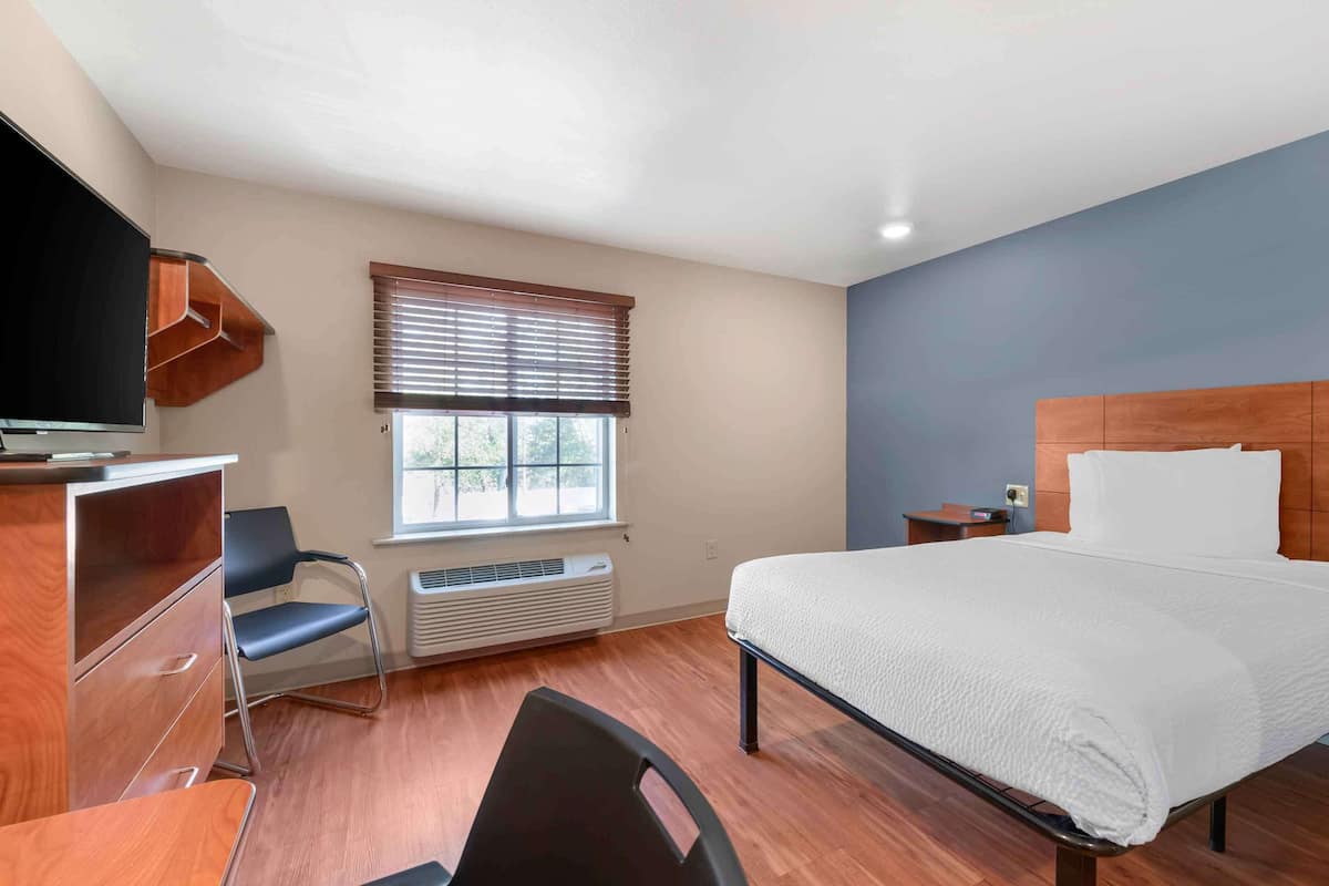 A hotel room with a bed, a desk, a chair, a television, a window with blinds, and a heater.