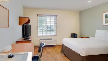 A hotel room with a bed, a desk with a television, a chair, a window with blinds, and an air conditioning unit.