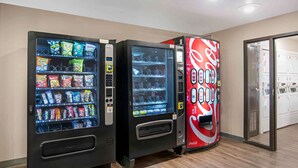 Drie automaten met snacks en dranken, waaronder een Coca-Cola automaat, en een deur die naar een kamer leidt.