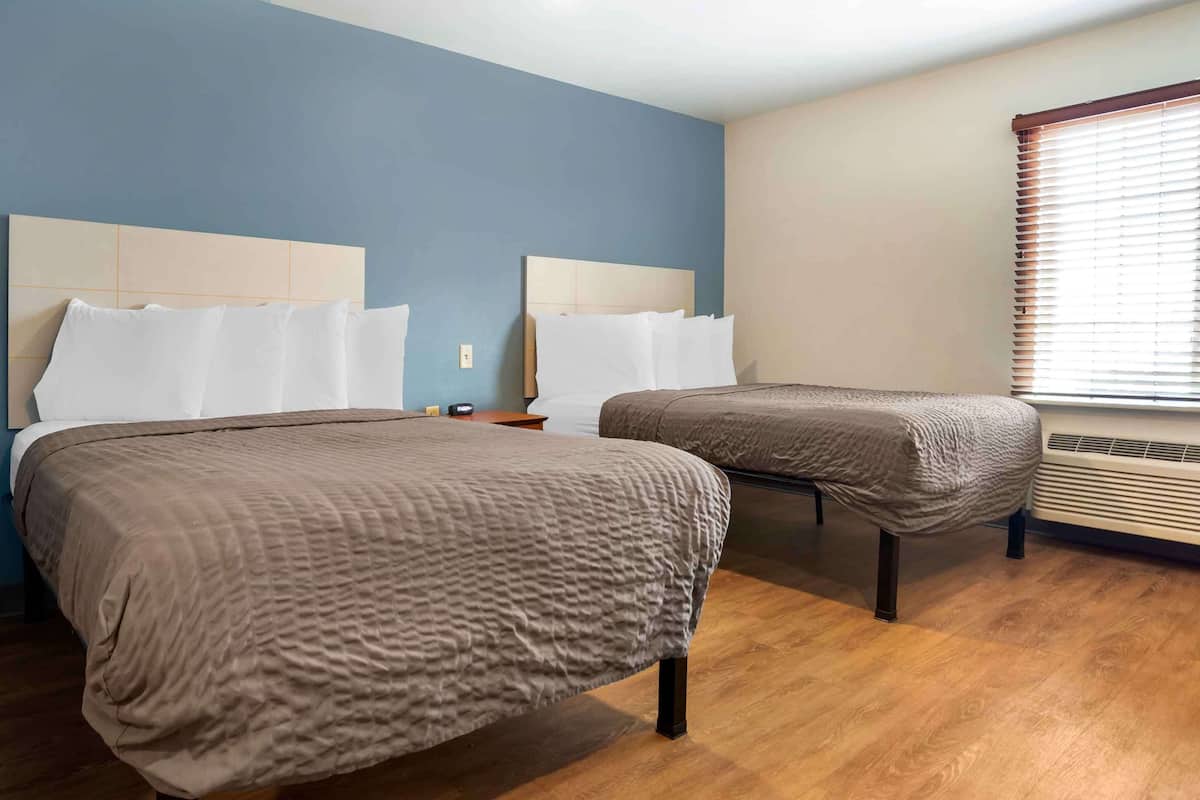A hotel room with two beds, a wooden floor, a window with blinds, and a wall heater.
