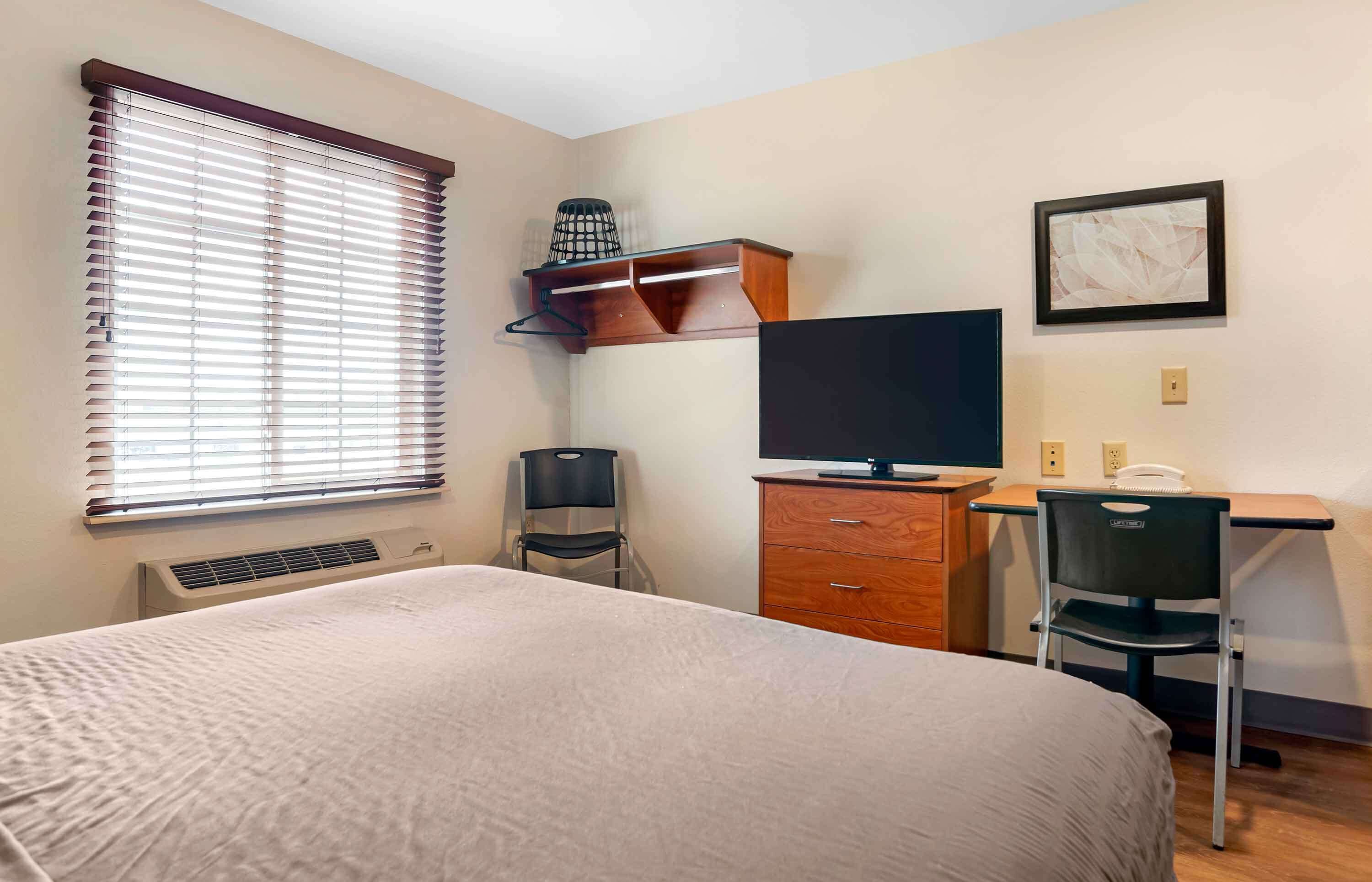 A hotel room with a bed, a desk with a chair, a television, a heater, and a window with blinds.