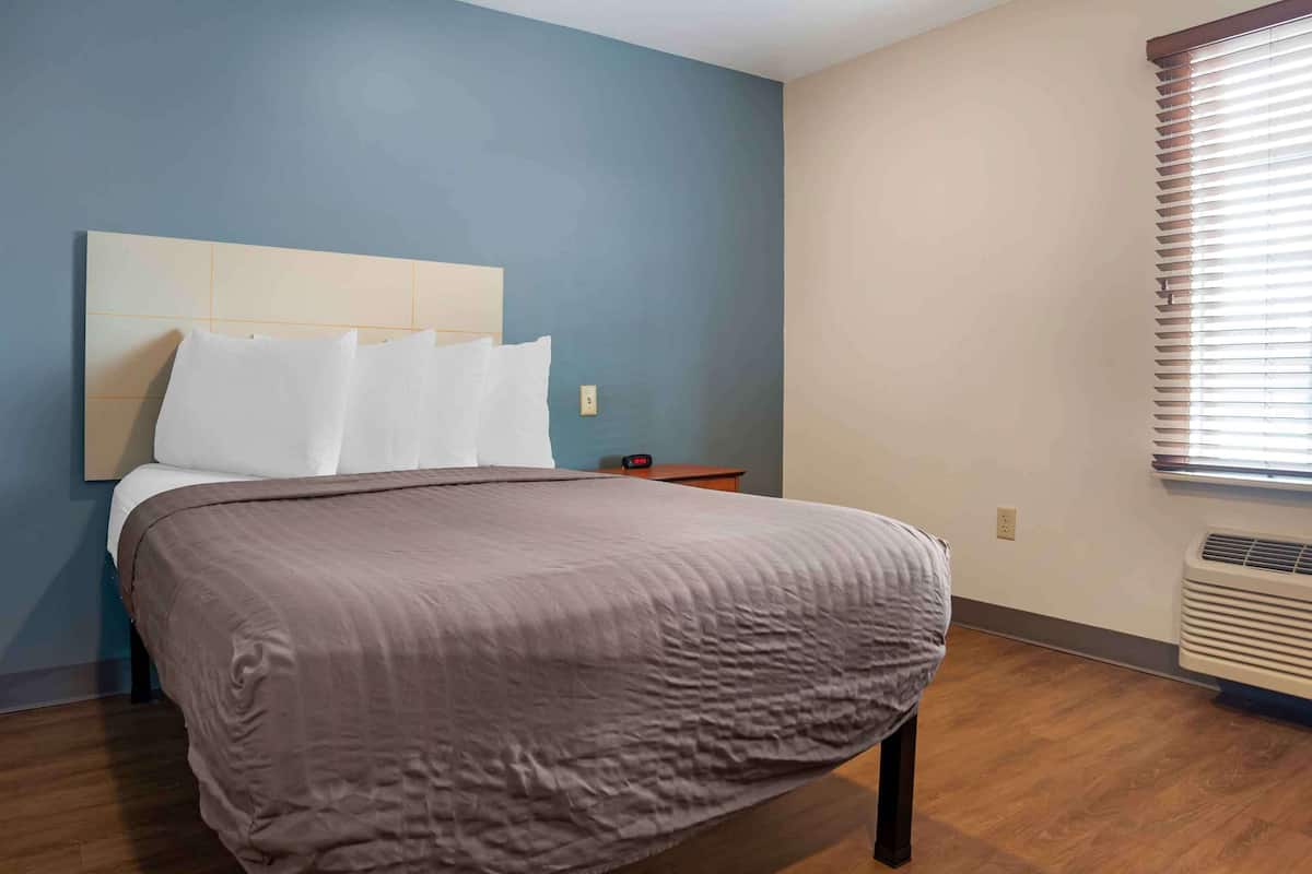 A neatly made bed with a headboard, a bedside table, and a window with blinds.