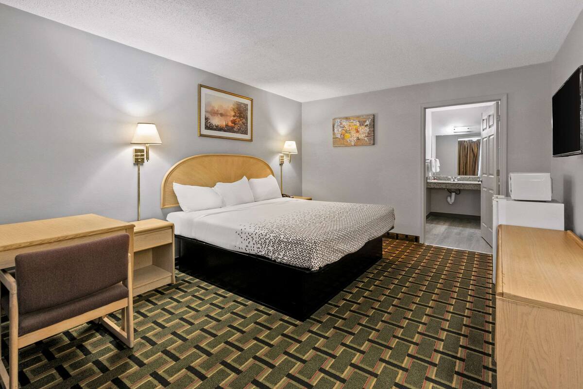 A hotel room with a bed, bedside table, chair, desk, and a bathroom visible through an open door.