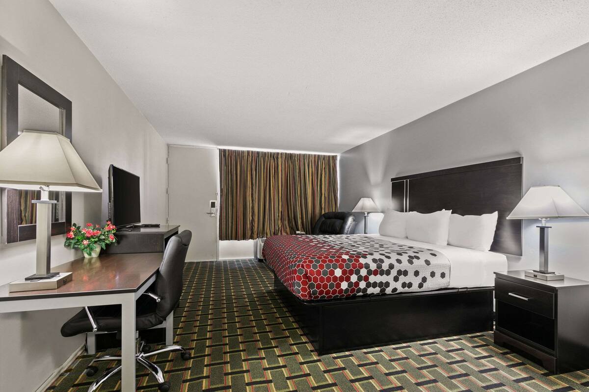 A hotel room with a bed, desk, chair, nightstand, lamp, and a view of the outside through curtains.