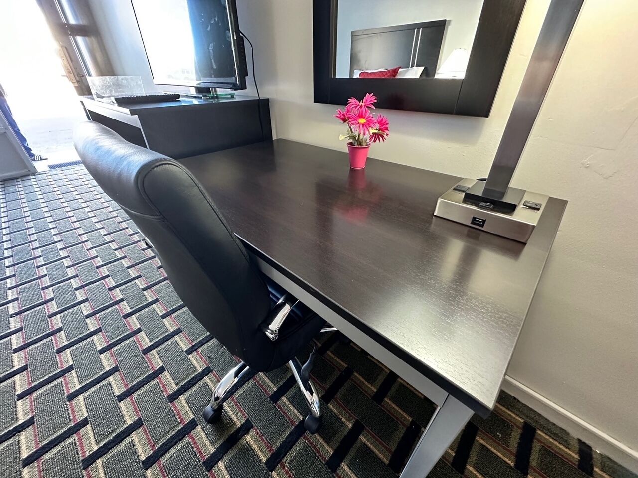 A modern hotel room with a large bed, a desk with a chair, a small table with a vase of flowers, and a large mirror on the wall.