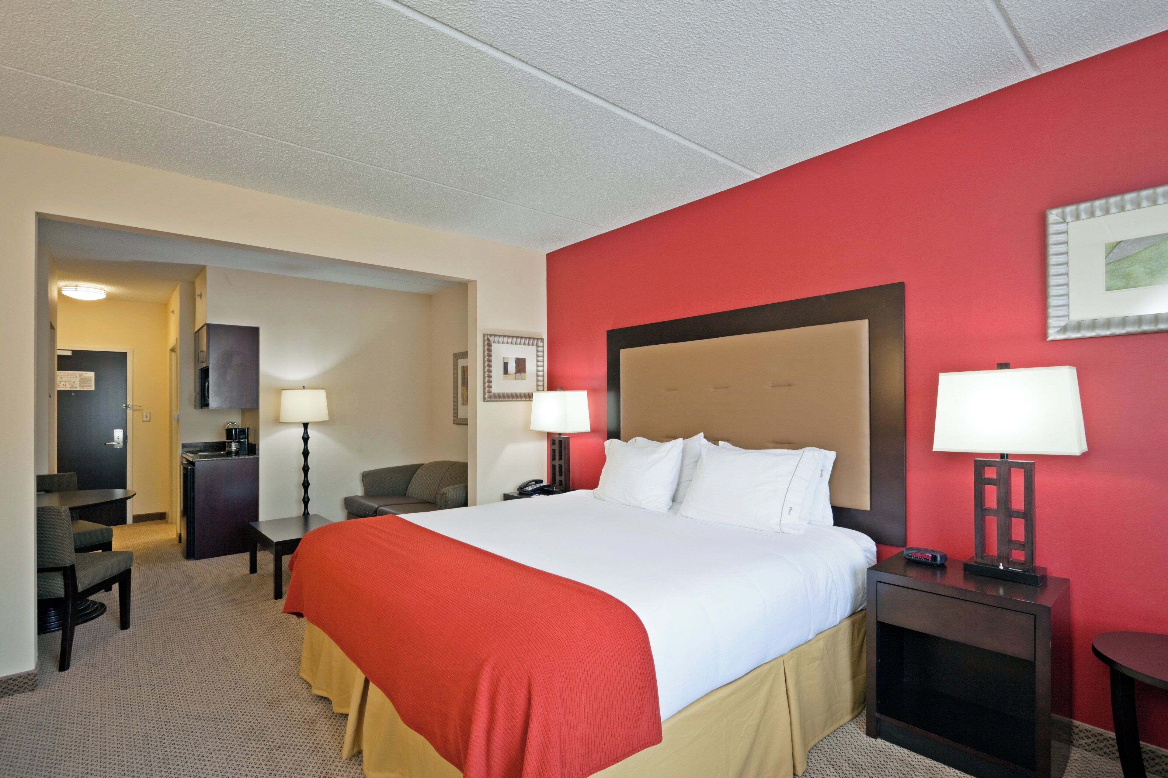 A hotel room with a large bed, a desk, a chair, a small kitchenette, and a red accent wall.