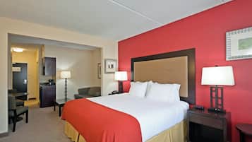 A hotel room with a large bed, a desk, a chair, a small kitchenette, and a red accent wall.