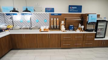 A breakfast bar with labeled sections for fruit, hot food, and cereal, along with a coffee machine and a refrigerator.