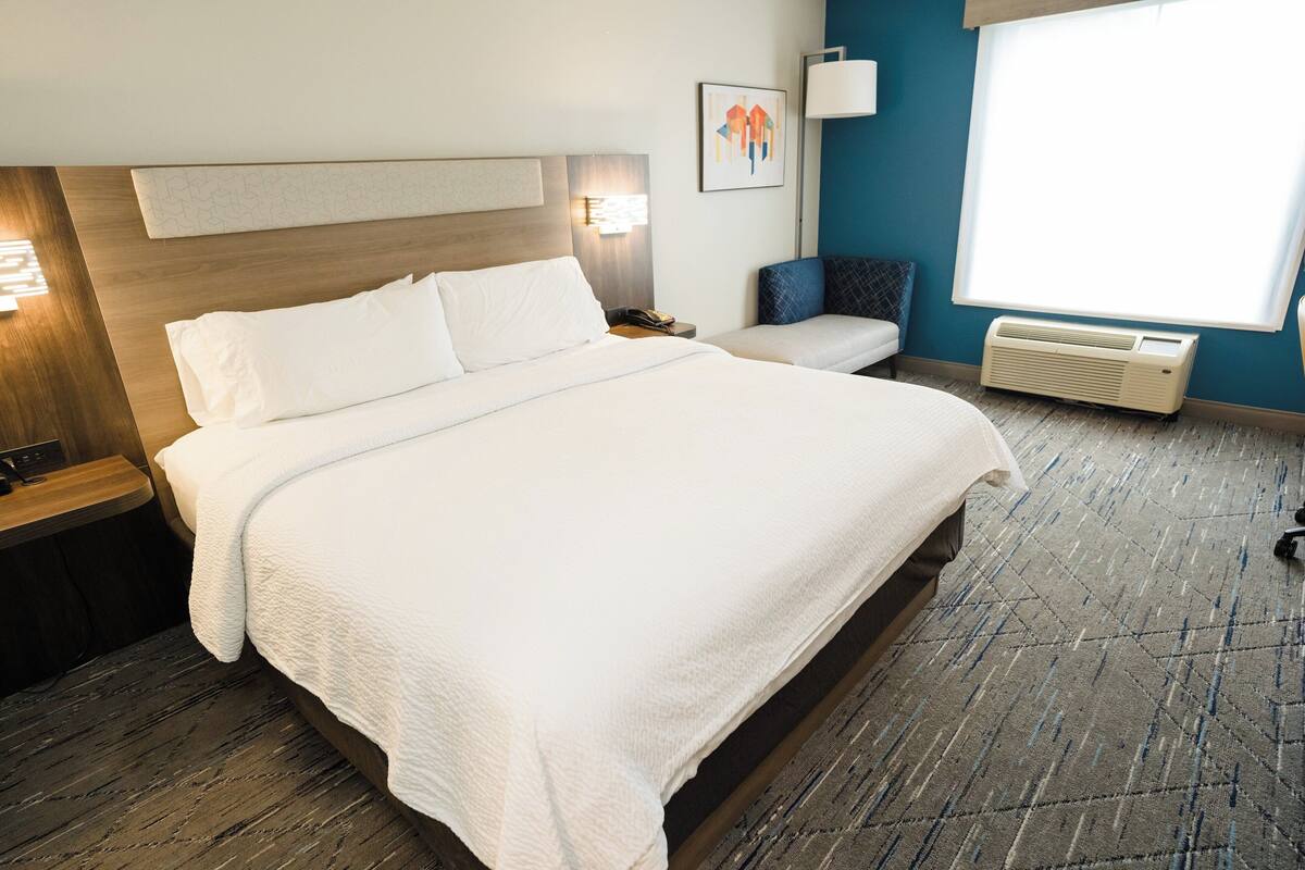 Una camera d'albergo con un letto grande, un comodino, una sedia, un condizionatore d'aria a muro e una finestra con tende.