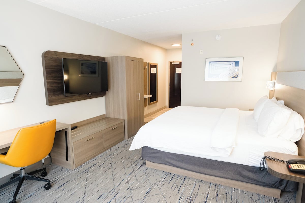Una camera d'hotel con un letto, una scrivania con una sedia, una televisione, un armadio e un comodino con una lampada.