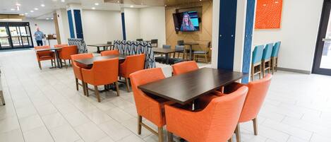 Un área de comedor moderna con sillas y mesas color naranja, una gran pantalla de televisión y un piso limpio con baldosas.