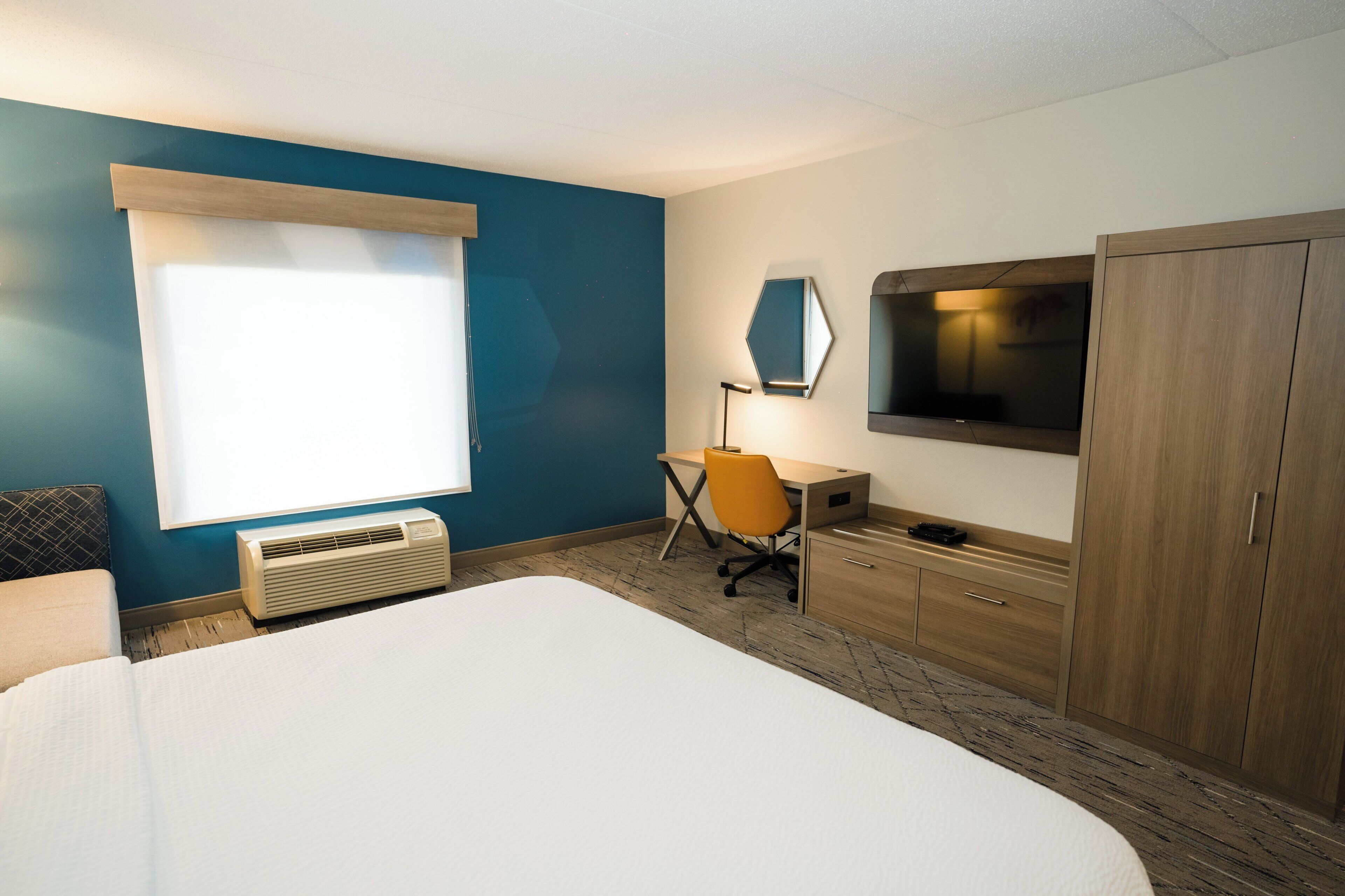 Habitación de hotel con cama, escritorio, silla, televisor, armario y ventana con persianas.