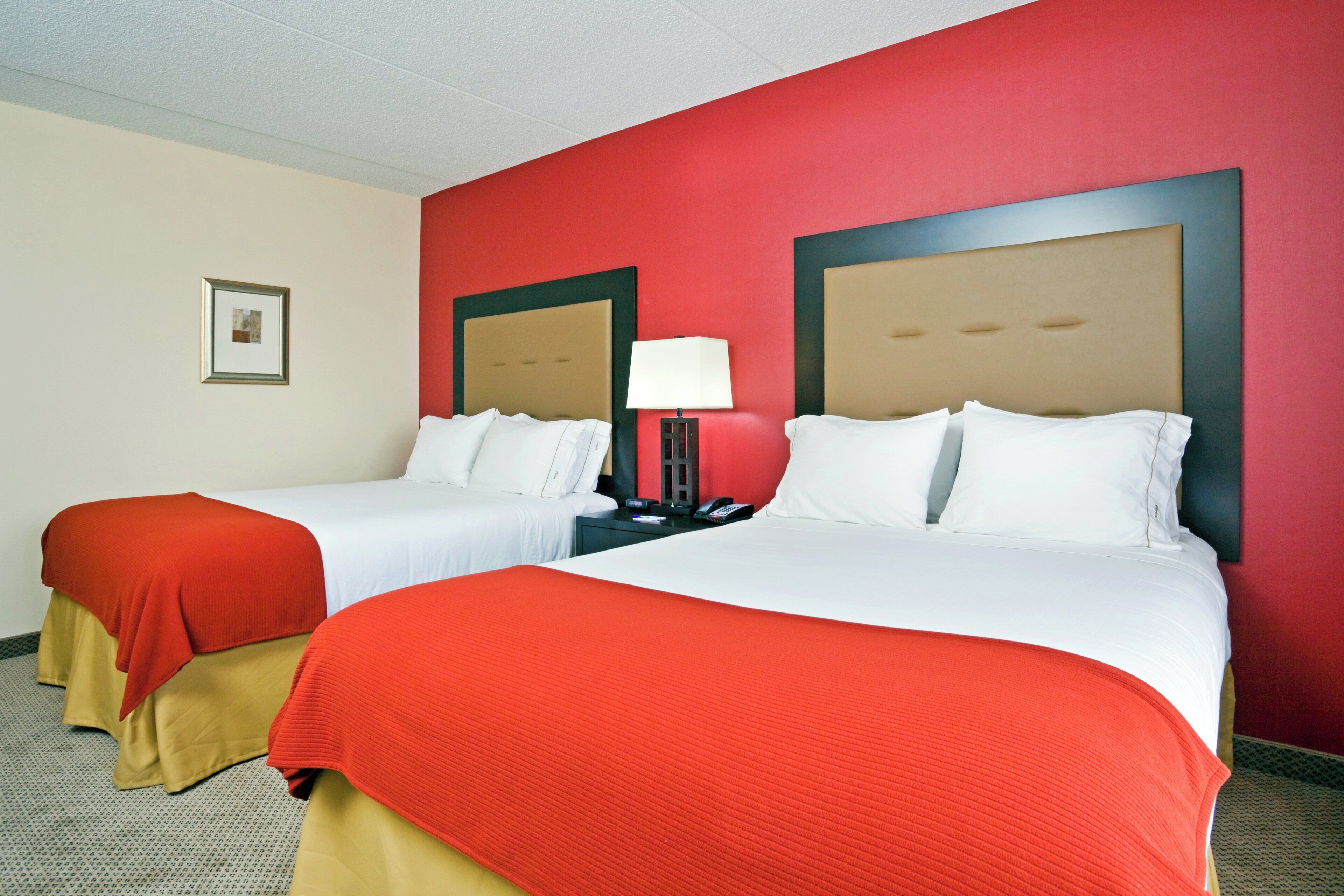 Habitación de hotel con dos camas, cada una con sábanas blancas y mantas rojas. Se observa un mesita de noche con una lámpara y un teléfono. Las paredes son rojas y hay un cuadro enmarcado en la pared.