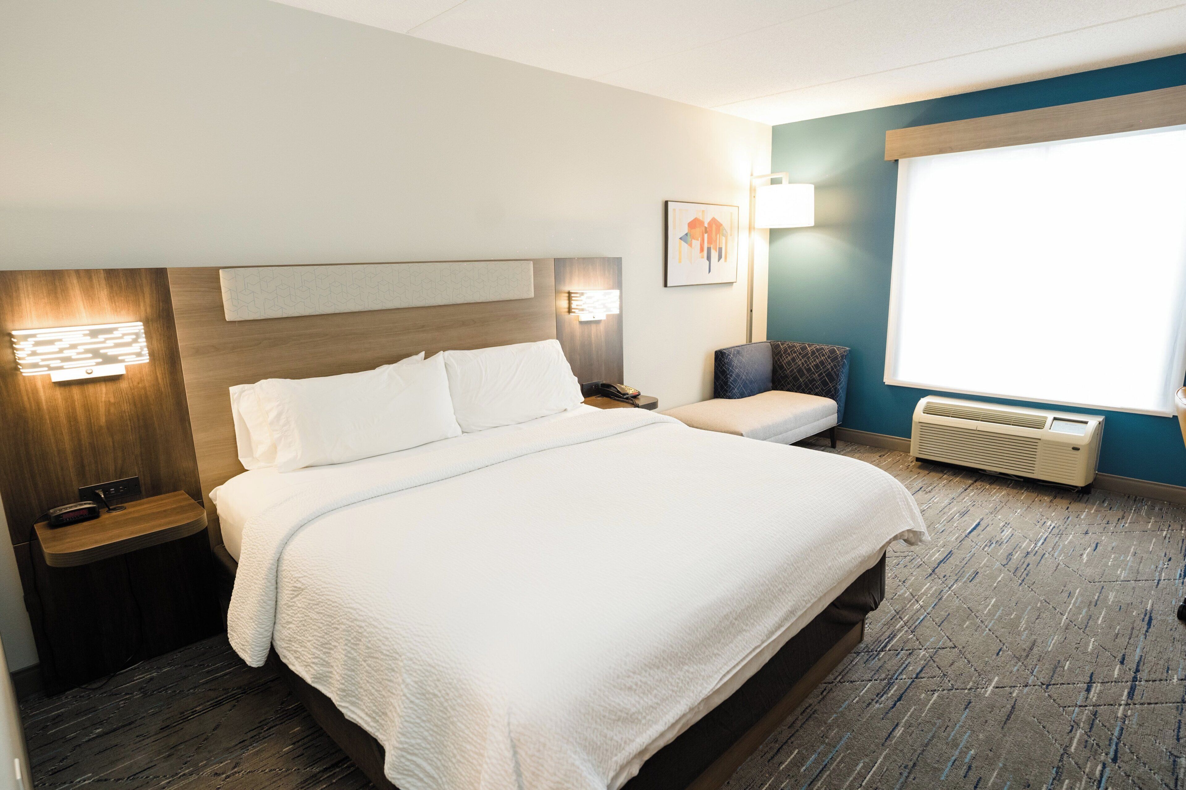 A hotel room with a large bed, bedside tables, a desk, a chair, a window, and a wall-mounted air conditioner.