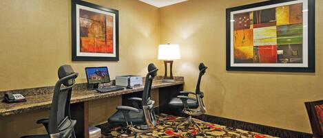 A hotel room with a desk, two chairs, a computer, a telephone, and two framed artworks on the walls.