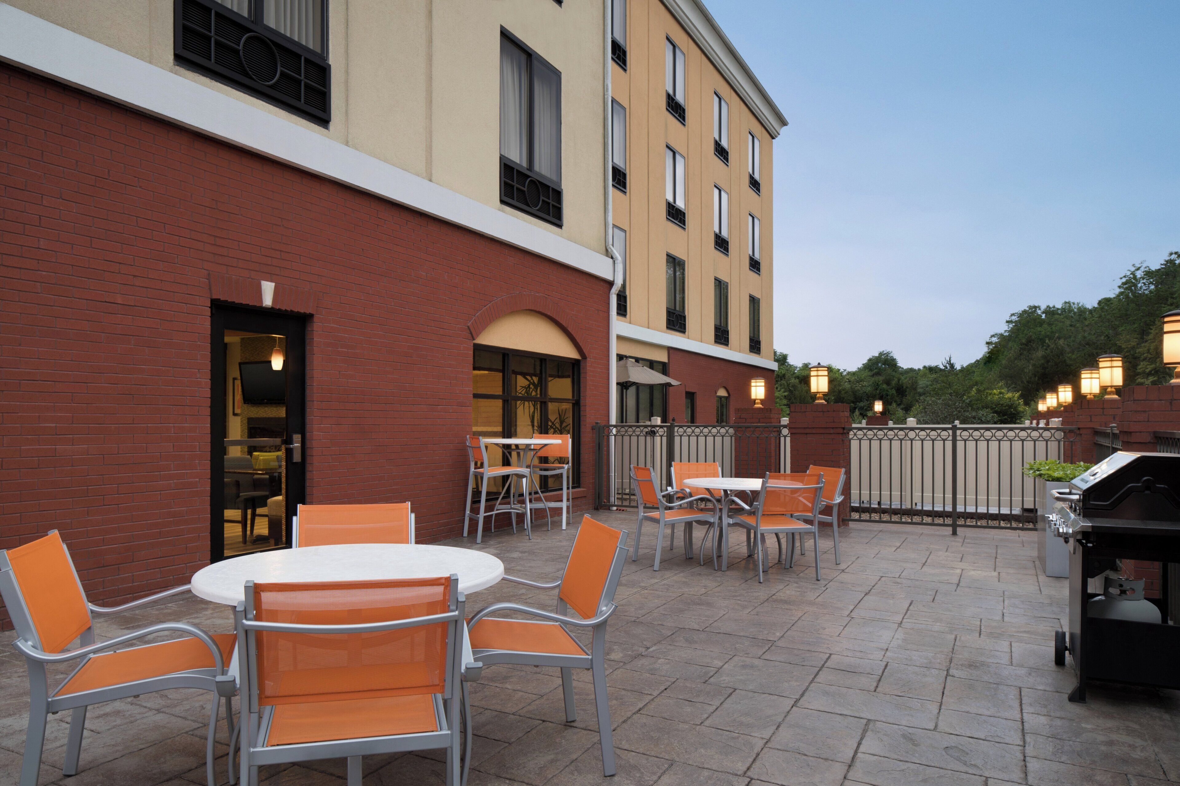 An outdoor patio area with tables and chairs, a grill, and a brick building in the background.