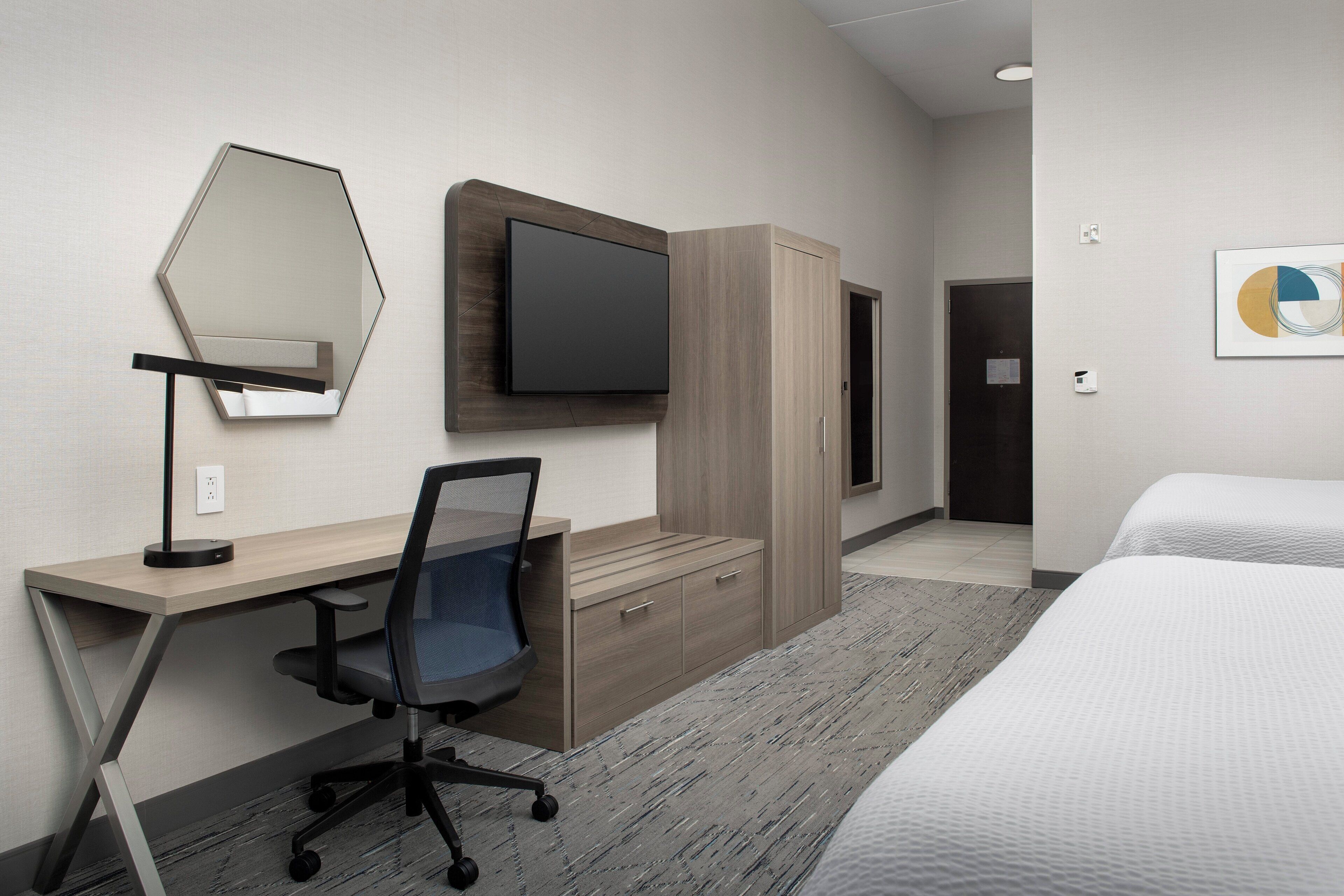 A hotel room with a desk, chair, bed, wardrobe, television, and artwork on the wall.