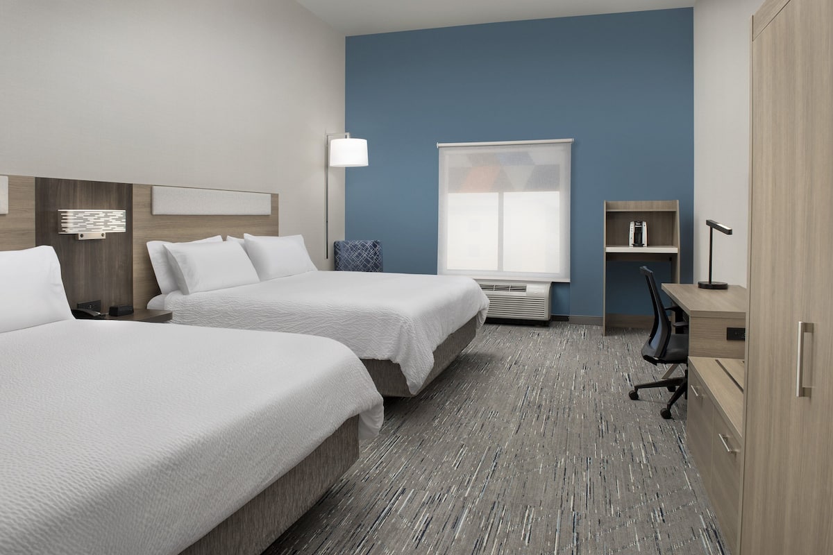 A hotel room with two beds, a desk, a chair, a lamp, a window, and a bookshelf.