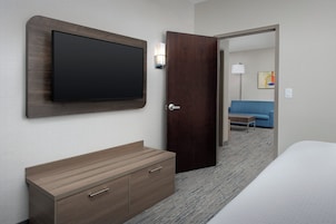 Habitación de hotel con televisión de pantalla plana montada en la pared, cómoda de madera, cama y puerta que conduce a otra habitación.