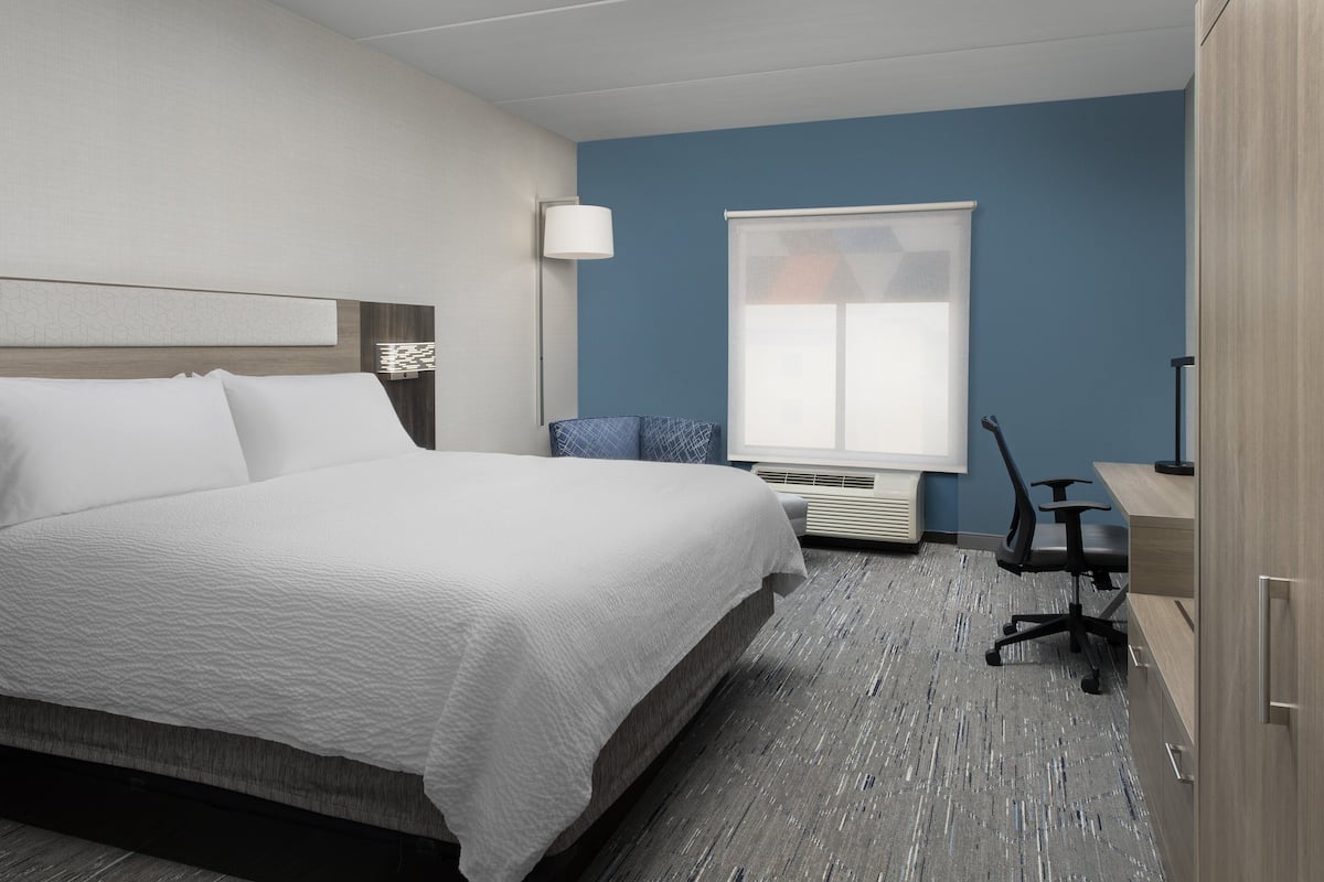 A hotel room with a large bed, a desk, a chair, a lamp, and a window with blinds.