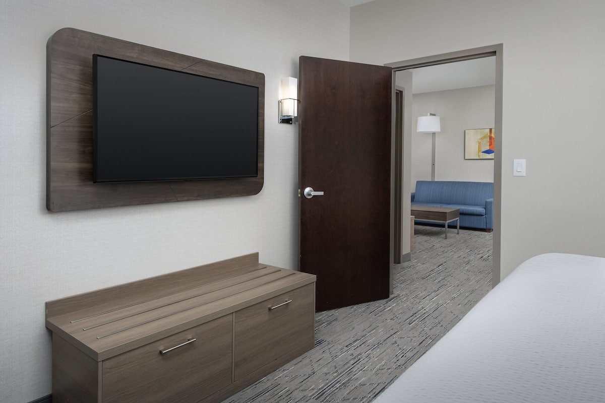 A hotel room with a flat-screen TV mounted on the wall, a wooden dresser, a bed, and a door leading to another room with a blue sofa and a lamp.