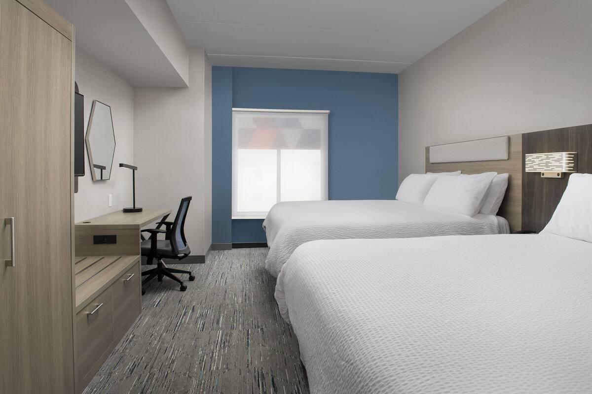 A hotel room with two beds, a desk with a chair, a wardrobe, and a large window with blinds.