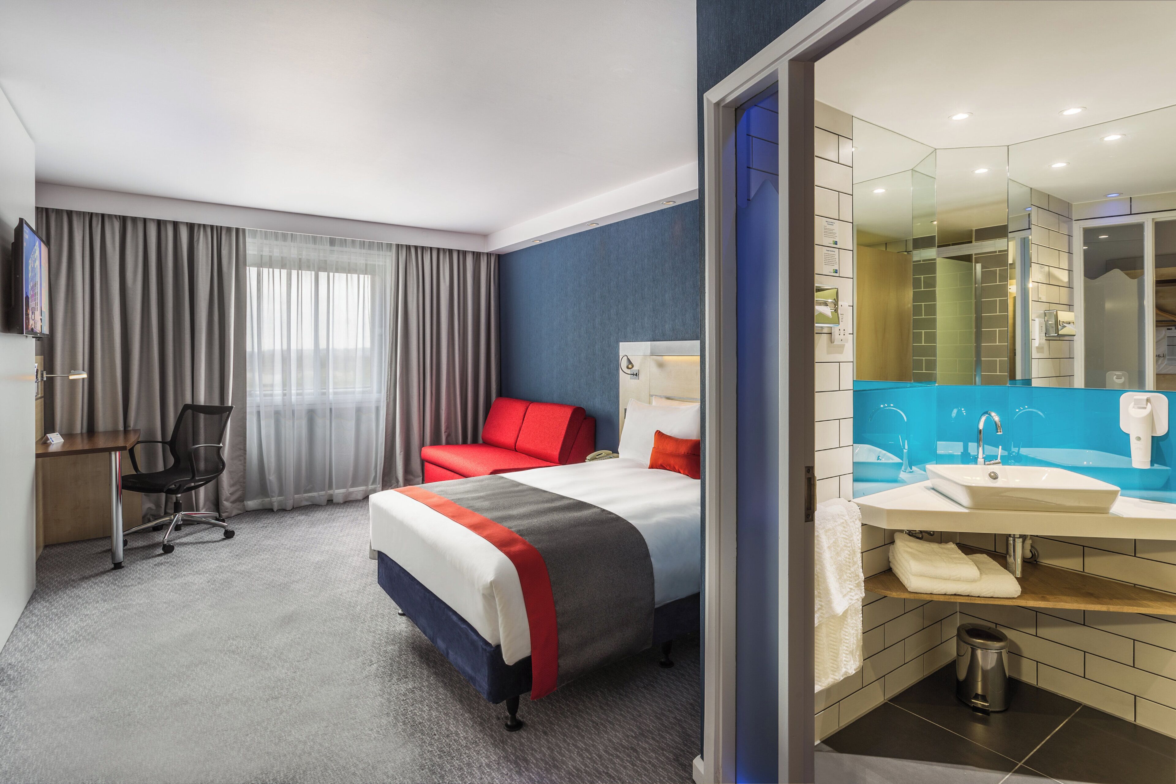 A hotel room with a bed, a desk, a chair, a TV, and a bathroom with a sink and mirror.