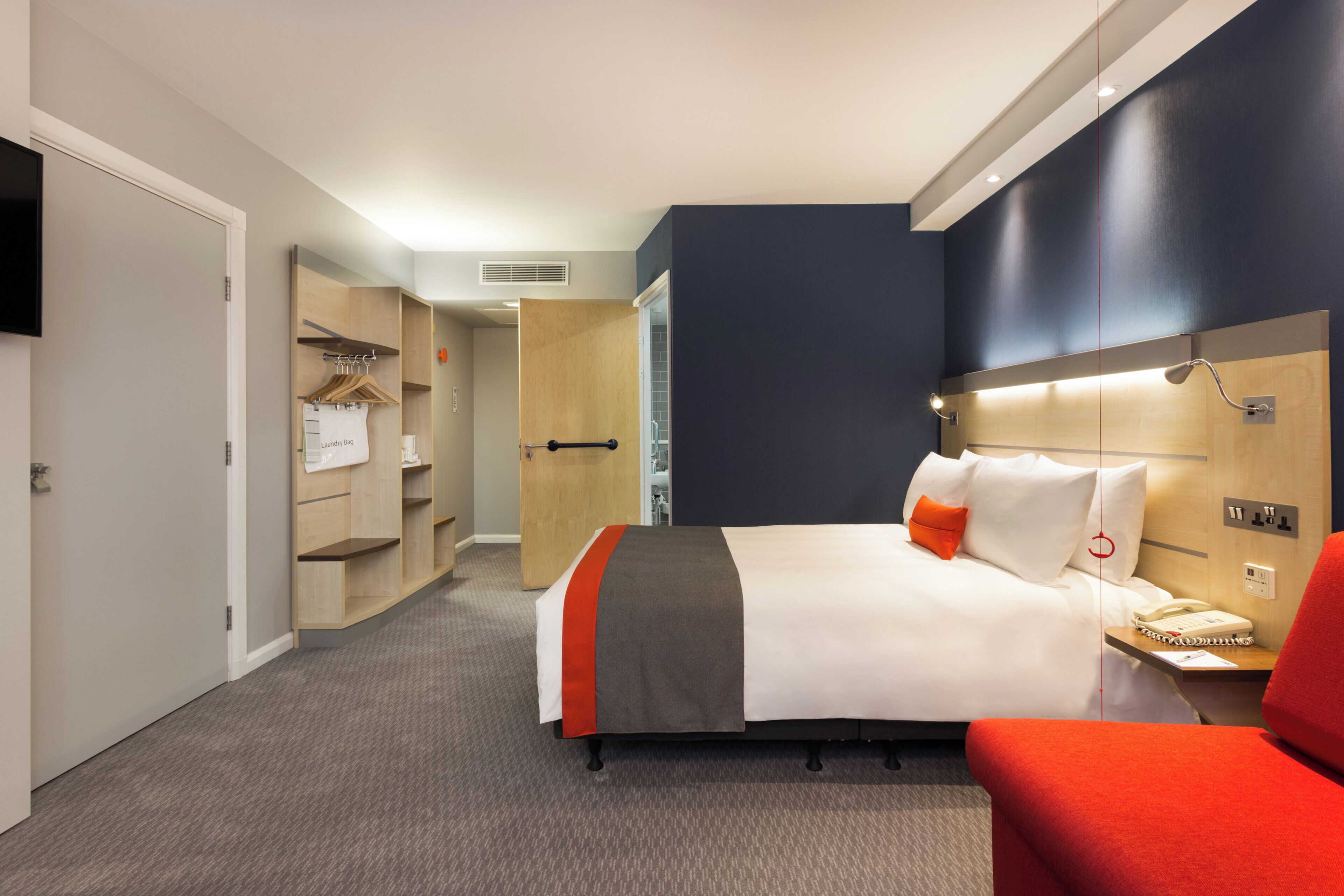 A hotel room with a bed, a bedside table with a phone, a red chair, a wardrobe, and a television on the wall.