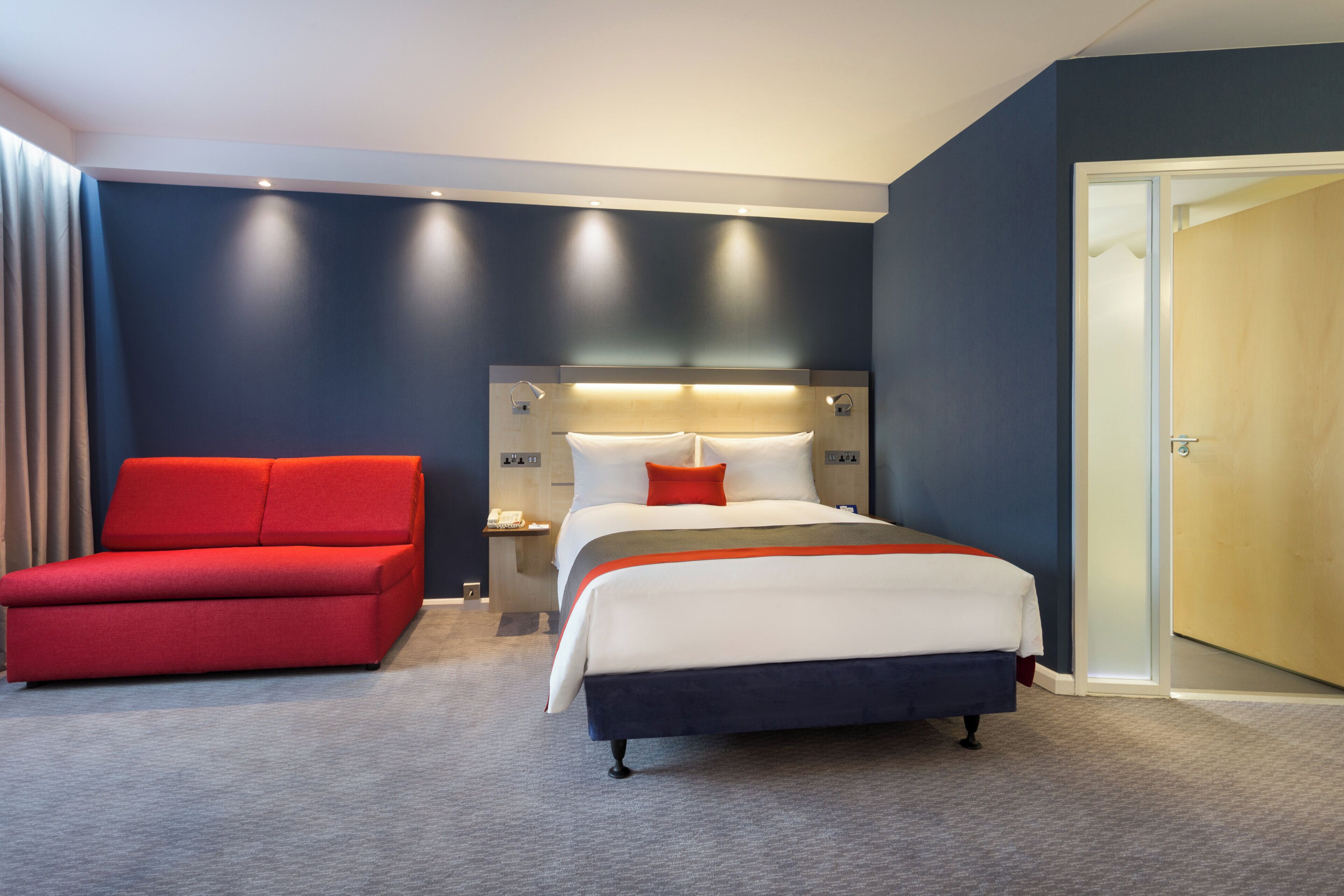 Una habitación de hotel con una cama, una silla roja, una mesita de noche y una puerta.