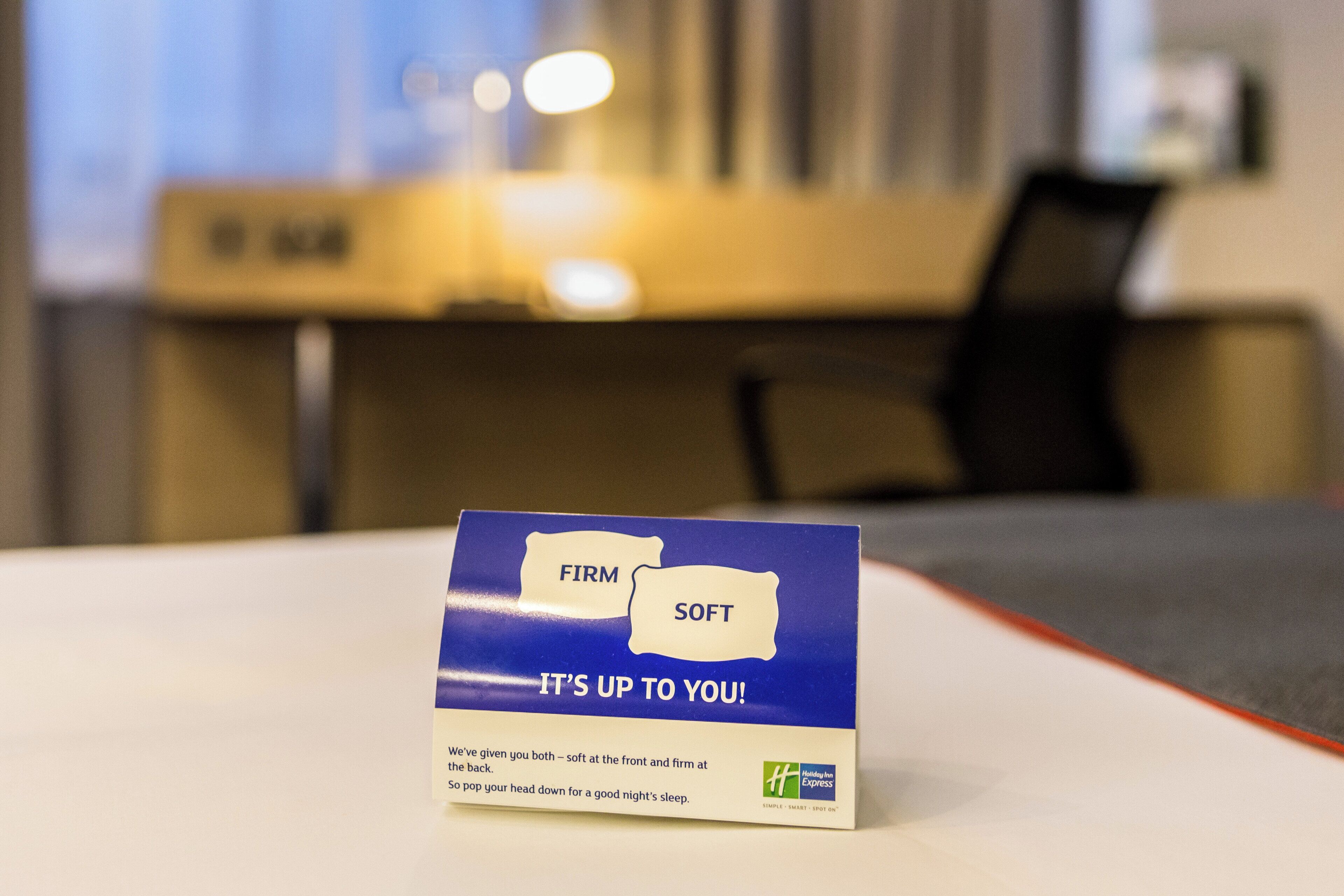 A hotel room with a bed, a desk, and a chair. There is a sign on the bed with the words 'FIRM SOFT' and 'IT'S UP TO YOU!' on it.