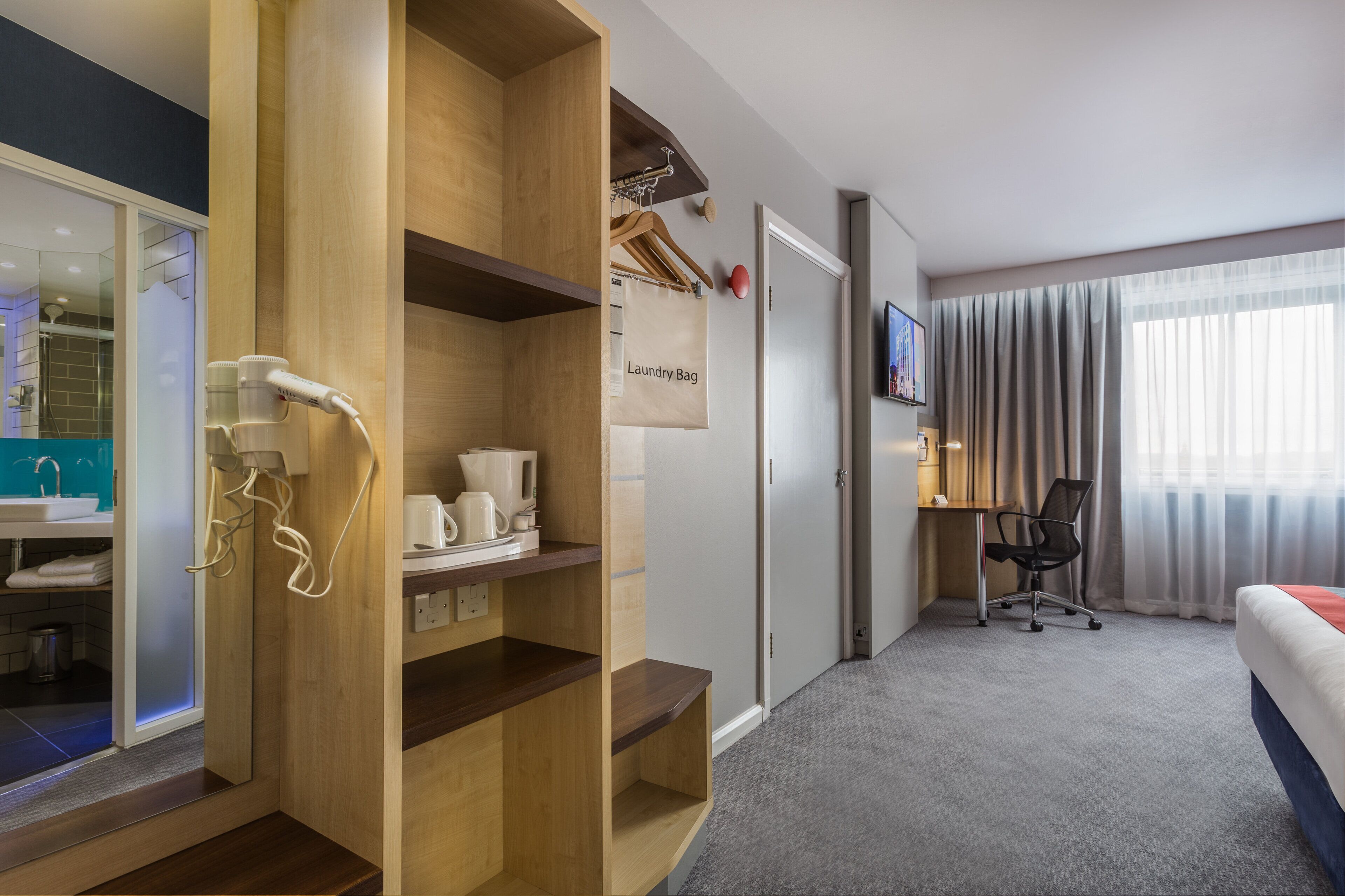 A hotel room with a wooden wardrobe, a coffee station, a desk with a chair, a bed, and a bathroom with a sink and mirror.