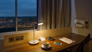 Ein Hotelzimmer mit Schreibtisch, einer Tasse Kaffee, einer Lampe und Blick auf die Stadt im Abendrot.