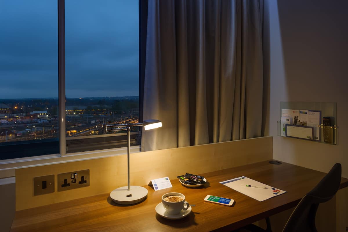 A hotel room with a desk, a cup of coffee, a lamp, and a view of the city at dusk.